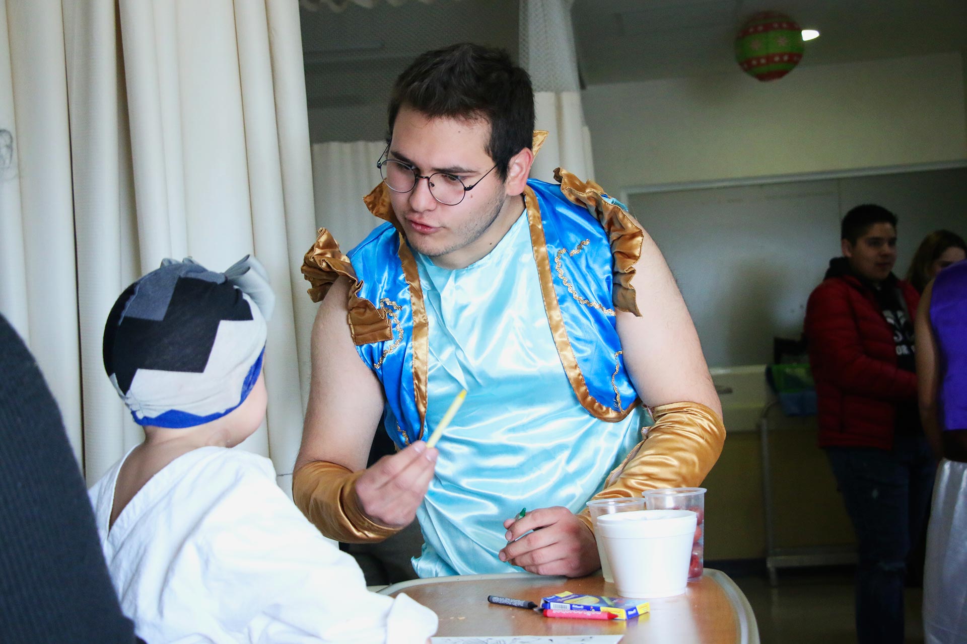 Aarón Vásquez, uno de los estudiantes del área de ingeniería, está caracterizado de el principe Aladdin juega con una niña y le ayuda a colorear un dibujo