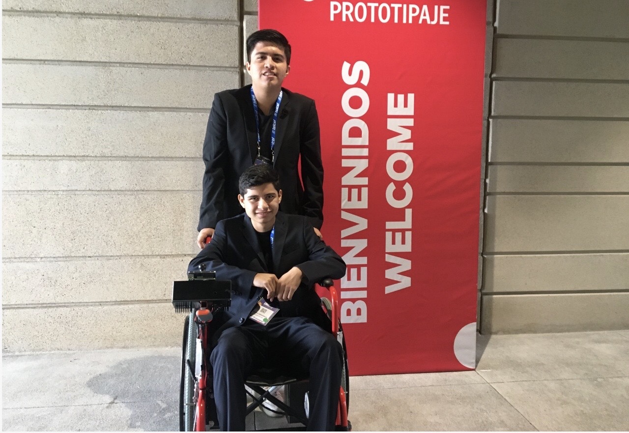 Daniel y Alexis con su prototipo de silla eléctrica frente a pancarta de congreso de prototipaje.