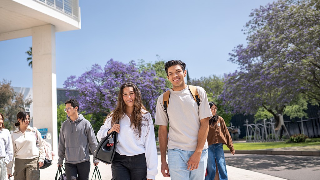 Estudiantes foráneos caminan por un campus al aire libre con mochilas, entre jardines y árboles con flores moradas, en un día despejado.