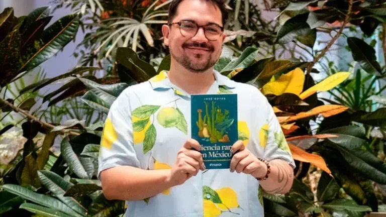 José Gotés (tarjeta 2): Hombre sonriente con lentes y camisa de estampado tropical sostiene un libro frente a plantas de hojas grandes
